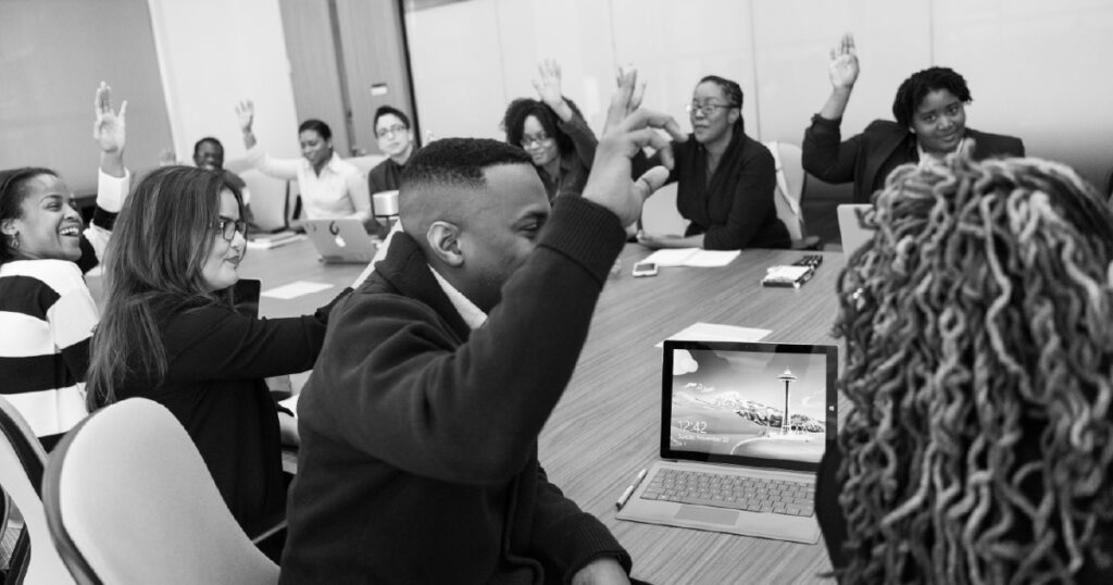 Adults around a table raise hands and smile during a presentation